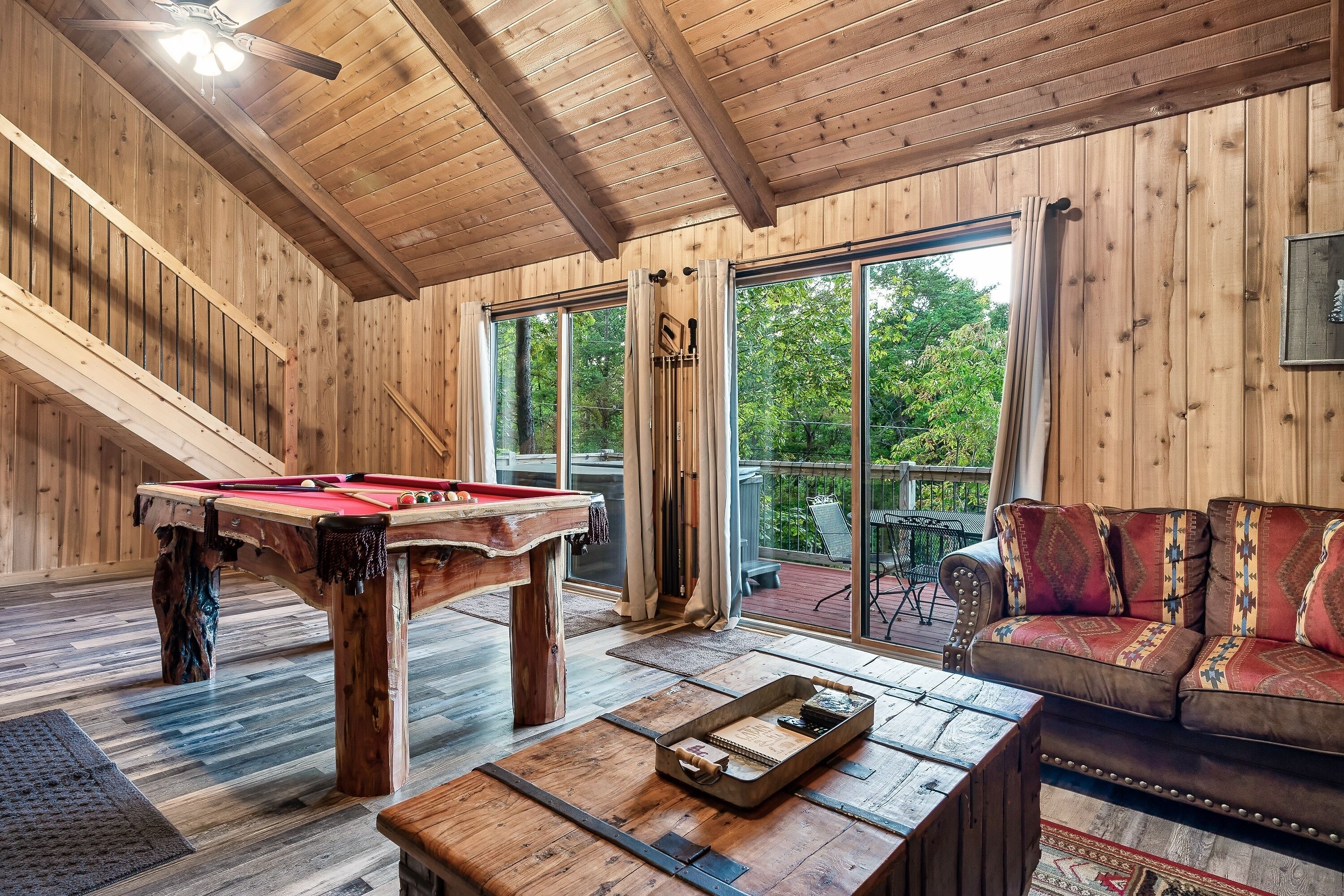 Ein Holzinterieur mit einem Billardtisch, einer Couch und einem Balkon mit Blick auf Bäume.