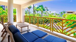 Un balcón con asientos acolchados azules, una mesa de madera y vistas a un paisaje tropical con palmeras y una playa.