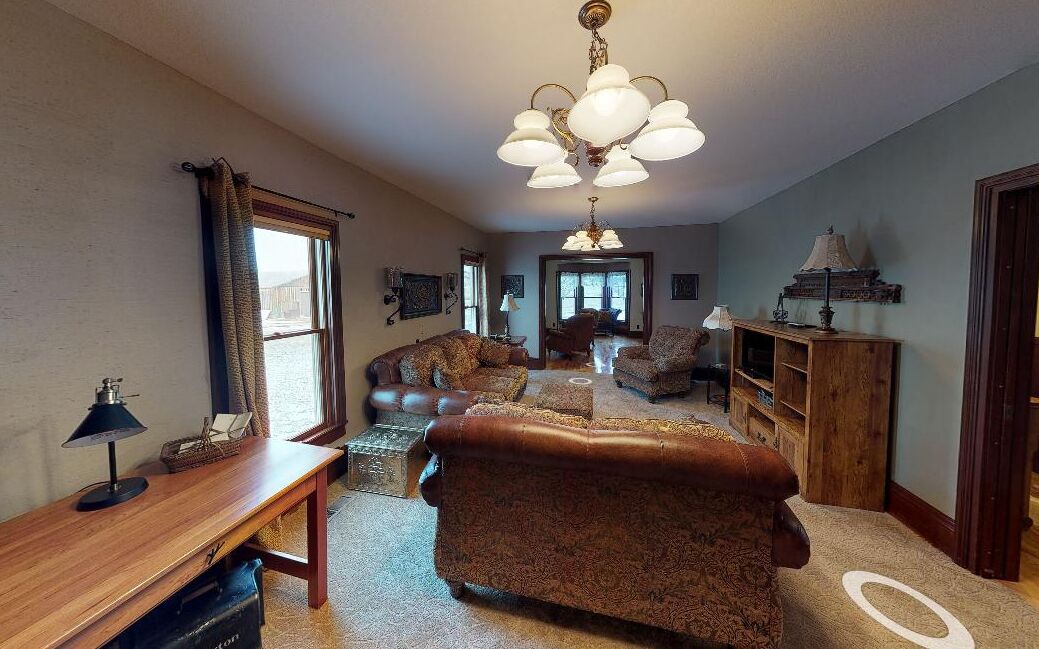 A spacious living room with a large chandelier, leather sofas, a wooden coffee table, and a bookshelf.