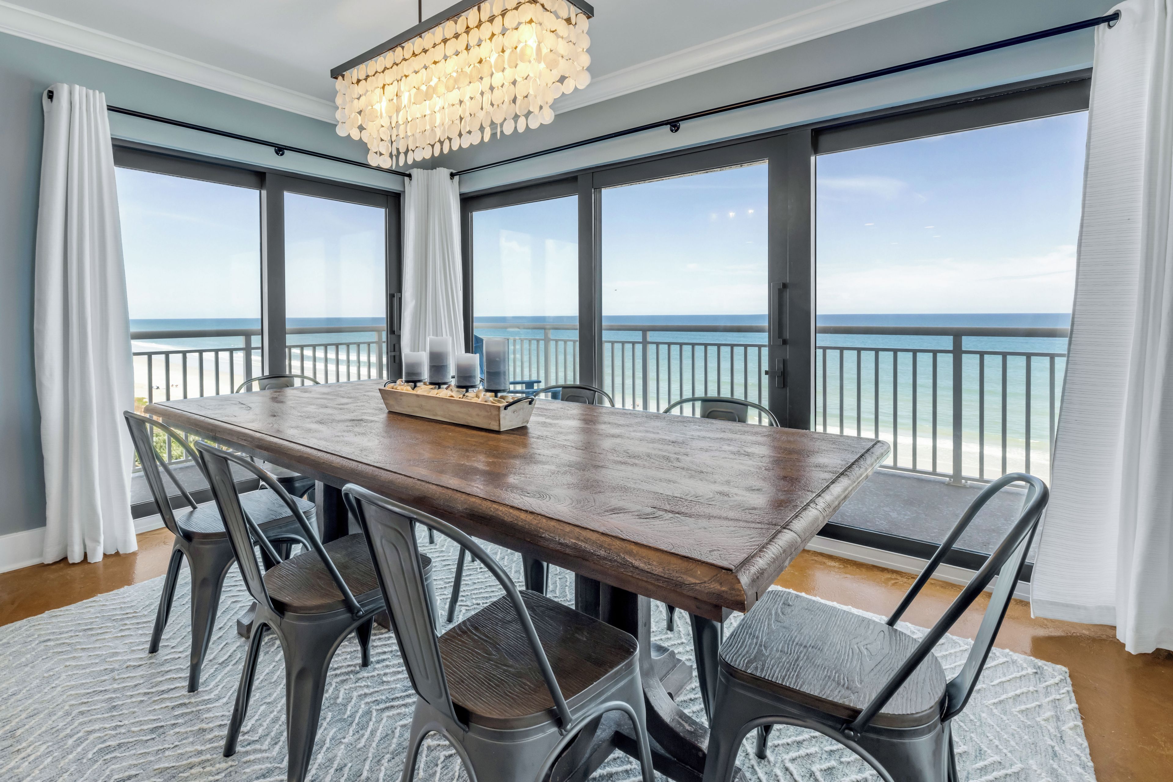 Comedor con mesa de madera, sillas de metal, una araña y vista al océano.