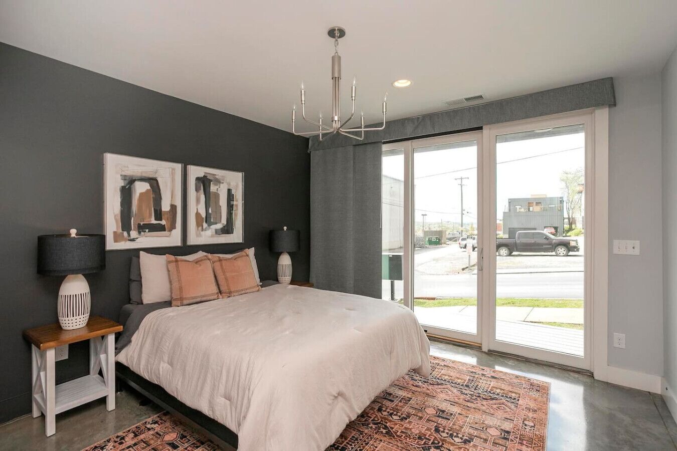 A bedroom with a large bed, two bedside tables, a chandelier, and a view of the street through a sliding door.