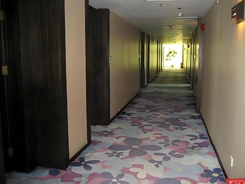 Un pasillo de hotel con alfombra floral, puertas de madera y un pasillo que conduce a una habitación luminosa.