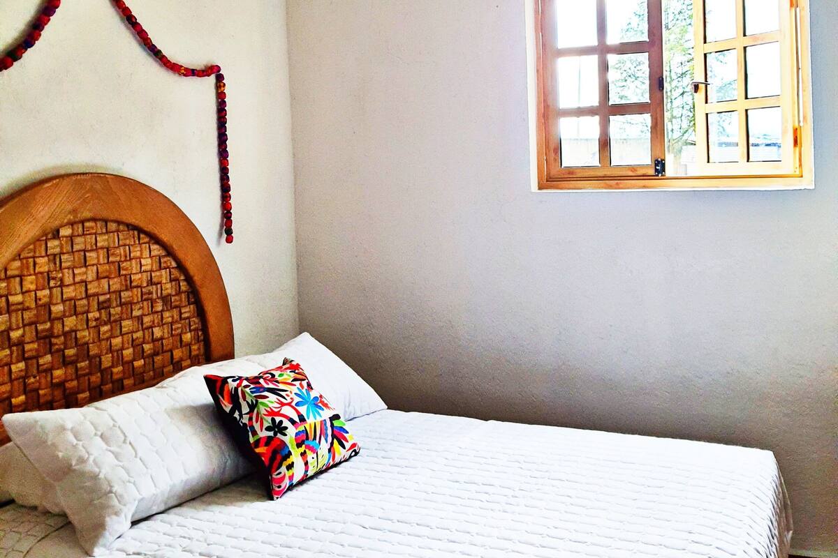 A bedroom with a bed, a colorful pillow, a woven headboard, a window with a view, and a hanging hot air balloon decoration.