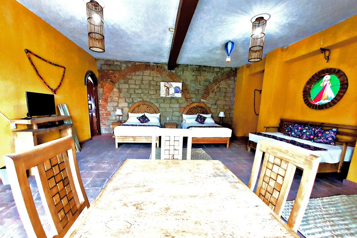 A room with two beds, a wooden table, and a bench. There are two lamps and a wall-mounted shelf with books.