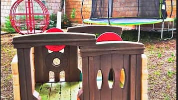 A wooden playhouse with a slide and a trampoline in the background.