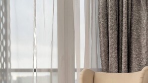 Una habitación con un ventanal grande, un sillón color beige, un reposapiés a juego y una mesita de madera.