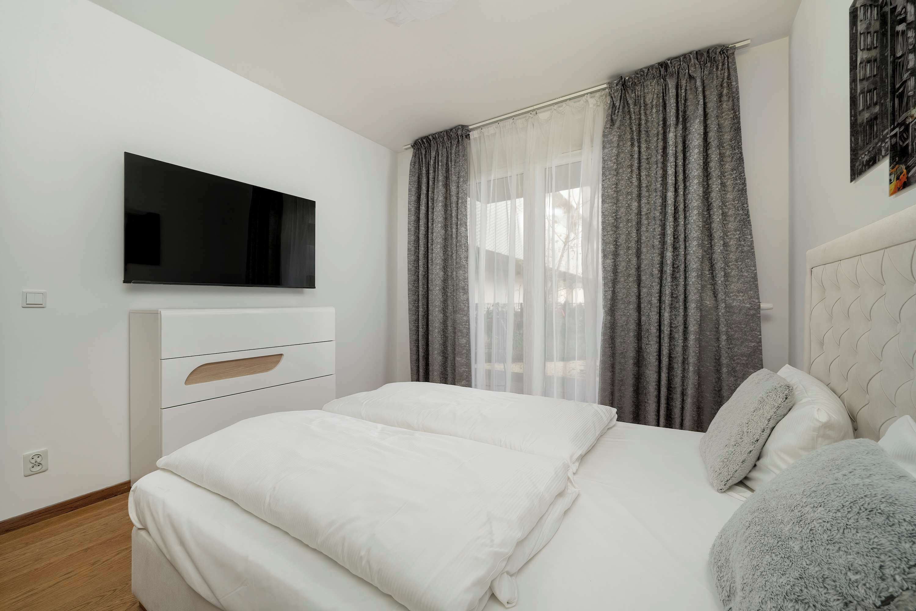 Ein Schlafzimmer mit einem Bett, einem an der Wand befestigten Fernseher, einem Fenster mit Vorhängen und einer weißen Kommode.