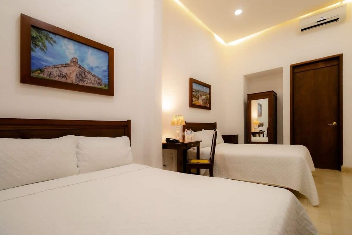Habitación de hotel con dos camas, un escritorio con lámpara, una silla, un espejo y dos cuadros en la pared.