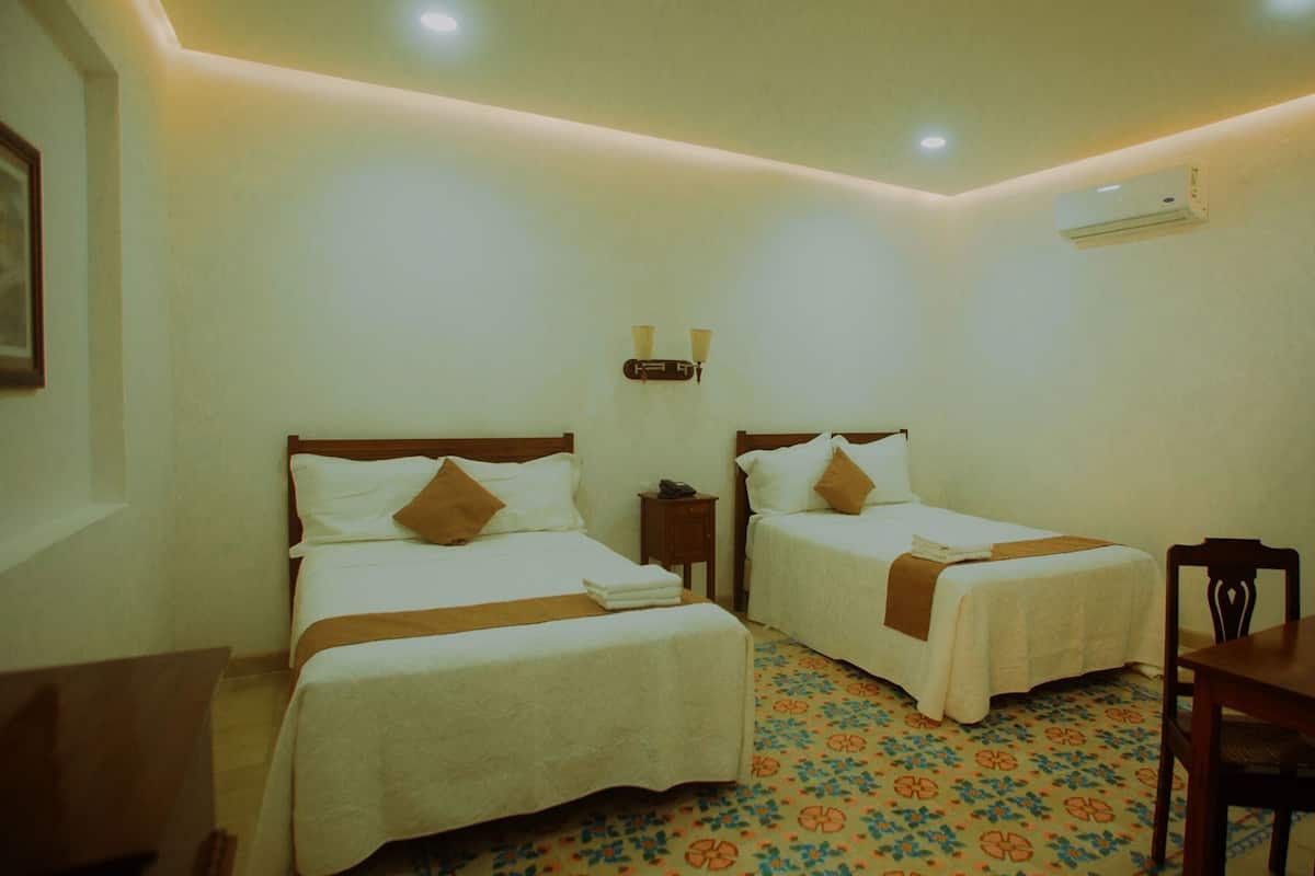 Habitación de hotel con dos camas, una mesita de noche con lámpara, aire acondicionado y alfombra estampada.