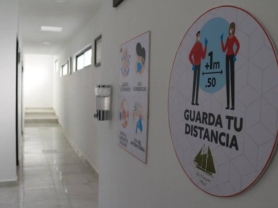 Un pasillo con un cartel que recomienda mantener una distancia de 1 metro, dispensador de gel hidroalcohólico y un letrero con el logotipo de un velero.