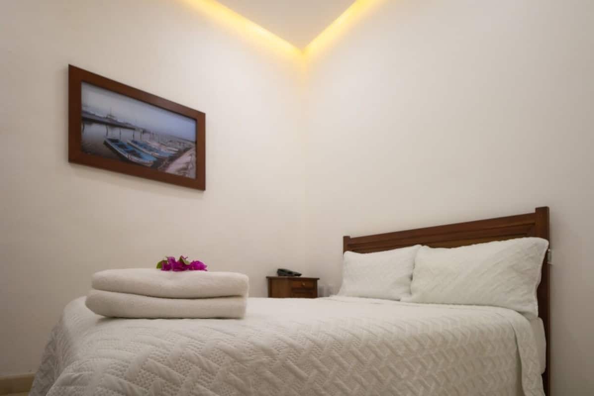 Una cama bien hecha con colcha blanca, cabecera de madera y una mesita de noche con un control remoto.