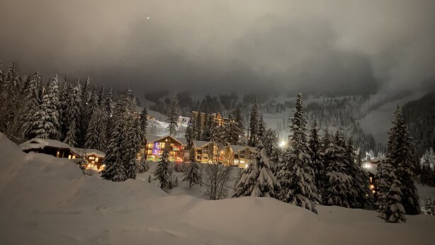 A snow-covered mountain village at night with illuminated buildings and snow-laden trees. - Condo C at The St. Bernard (Sandpoint)