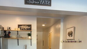 A modern kitchen with wooden cabinets, a stainless steel refrigerator, and a countertop. There is a hallway leading to a door and a small seating area with a lamp. - Condo C at The St. Bernard (Sandpoint)