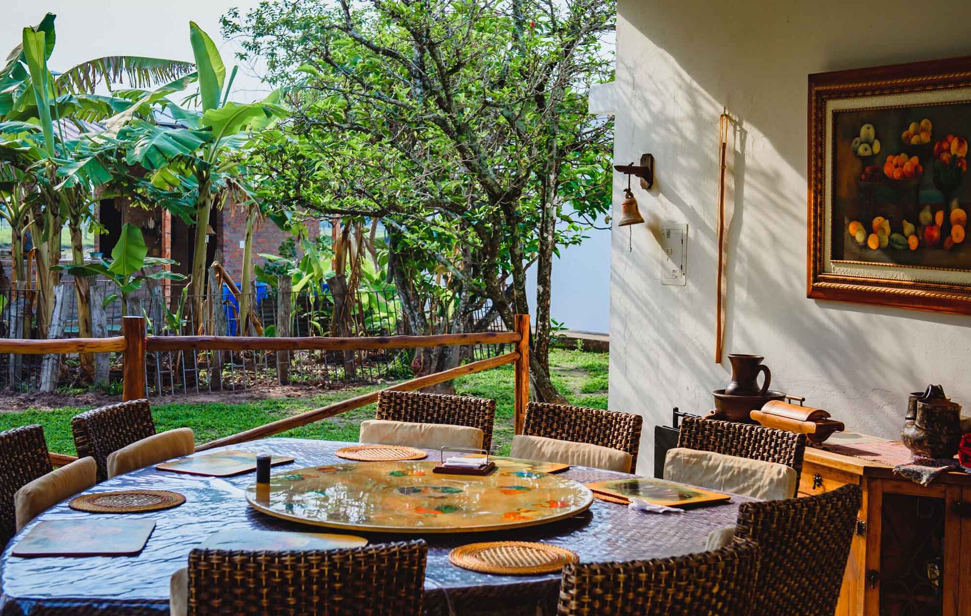 An outdoor dining area with wicker furniture and a glass-top table, surrounded by lush greenery and a view of the sea.