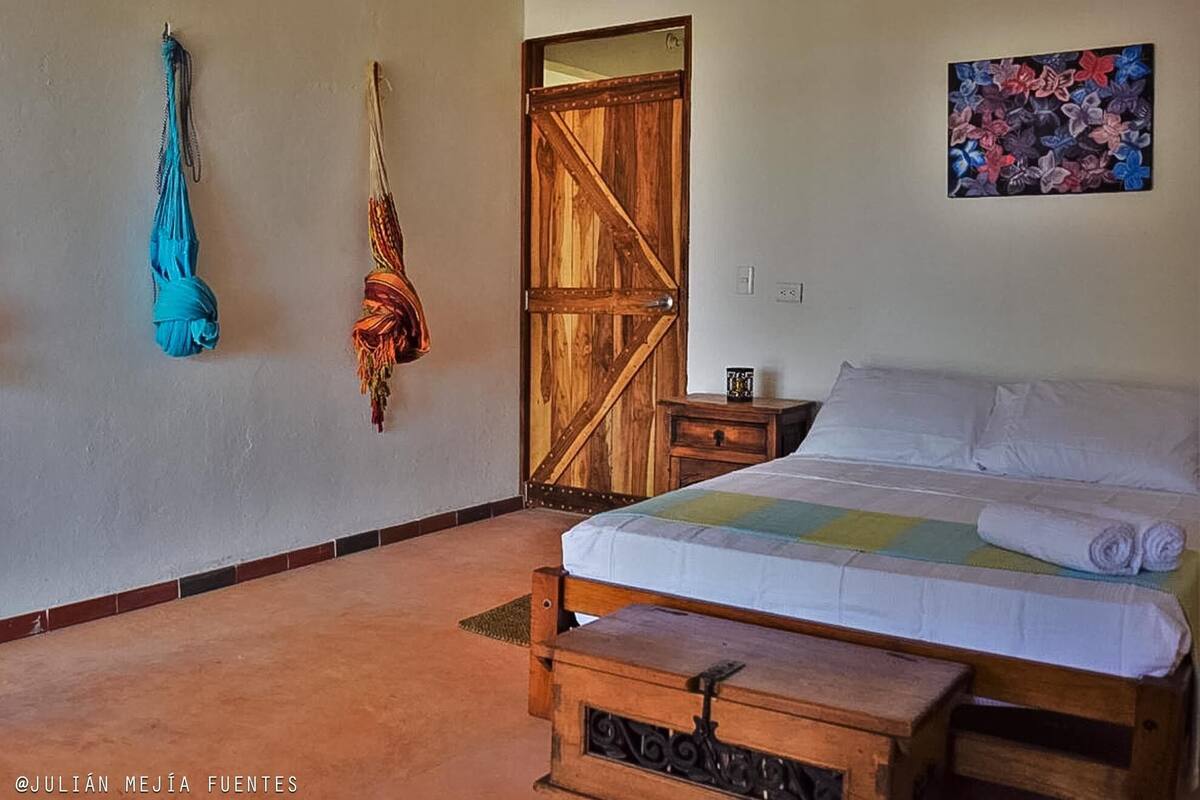 Un dormitorio con una cama de madera, una mesita de noche, un baúl de madera, una puerta y una pared con un cuadro colorido.