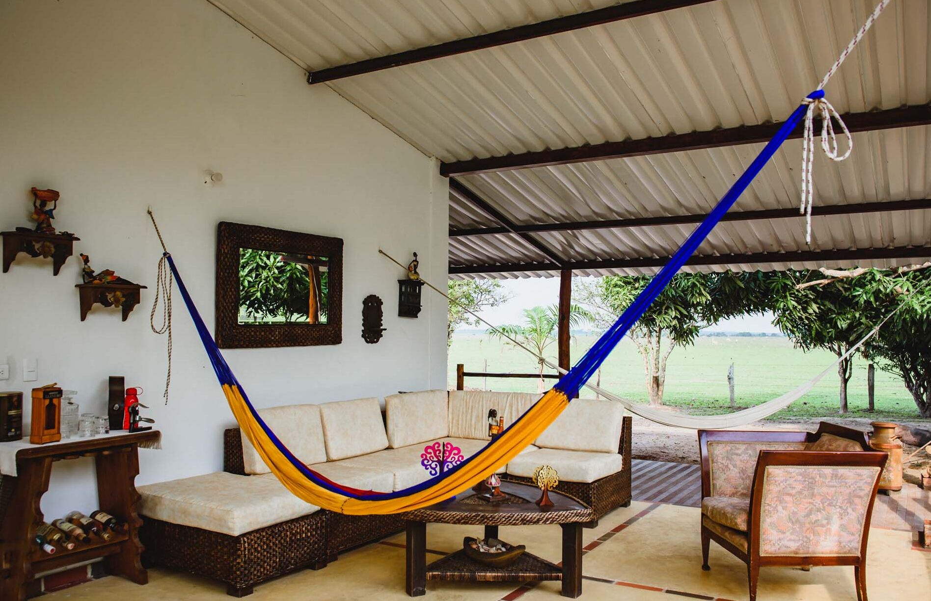 A hammock suspended between two wooden beams in a room with a white wall, a wicker sofa, a small table with bottles, and a view of a green landscape.