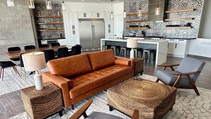 Ein modernes Wohnzimmer mit einer Couch, Sesseln und einem Couchtisch. Es gibt eine Küchenzeile mit einem Kühlschrank sowie einen Esstisch mit Stühlen.