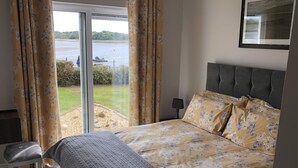 Un dormitorio con cama, un banco, una ventana con cortinas, una lámpara y vista a un cuerpo de agua.