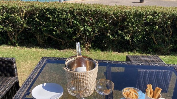 Mesa preparada con comida, una botella y vasos, con vista a un cuerpo de agua, una cerca y follaje al fondo.