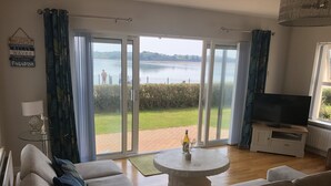 Una sala de estar con un sofá, un televisor sobre un mueble bajo, una mesa de vidrio con una botella, y vista a un lago a través de puertas correderas grandes.