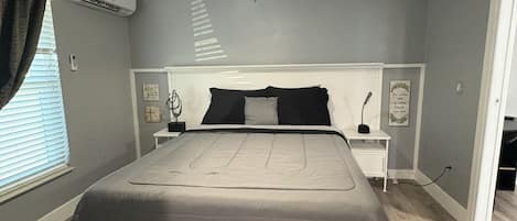 A bedroom with a bed, a ceiling fan, an air conditioning unit, a nightstand, and a bedside table.