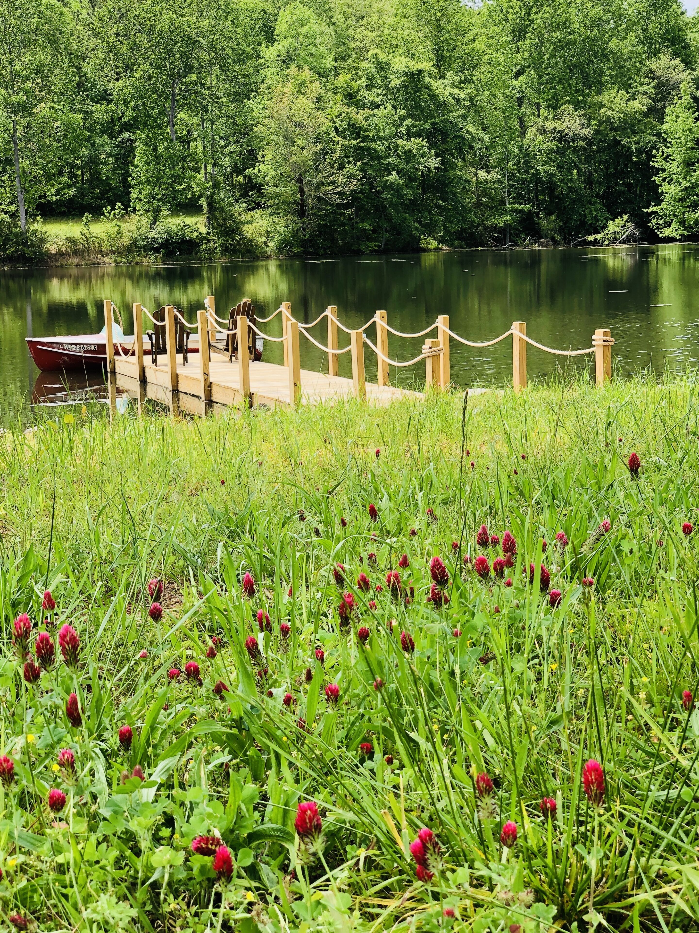 A wooden dock extending into a calm lake, surrounded by lush greenery and red flowers.