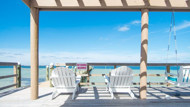 Eine Holzterrasse mit zwei weißen Adirondack-Stühlen, die auf einen Steg zeigen, an dem ein Schild mit der Aufschrift 'NO DIVING ALLOWED' angebracht ist.