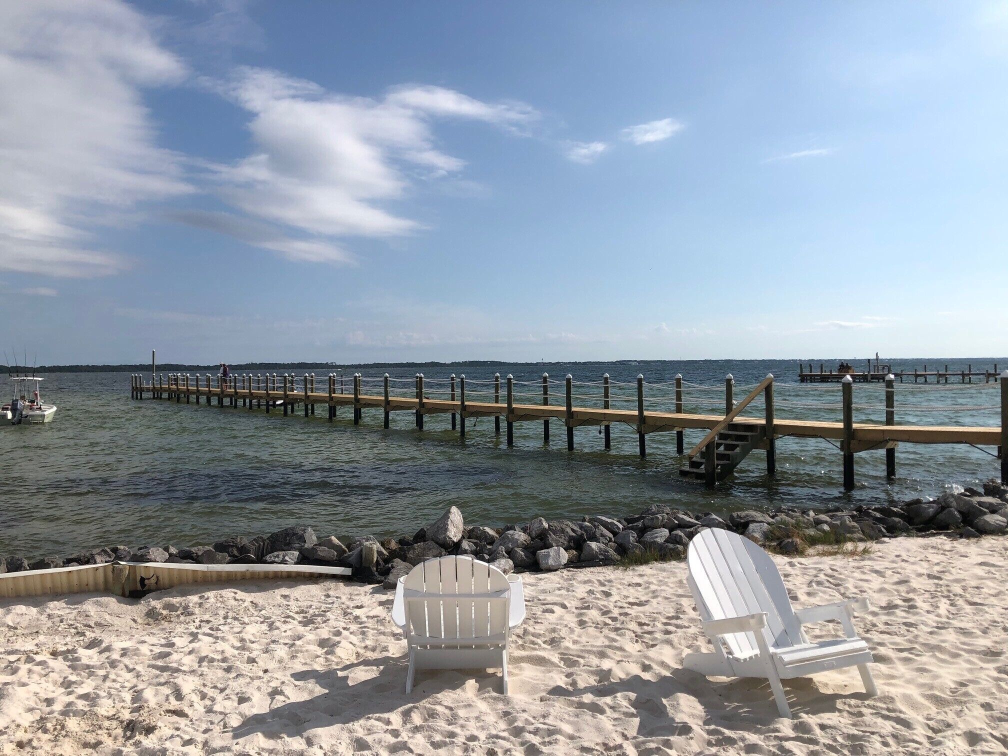 Una playa con dos sillas Adirondack blancas, un muelle de madera que se adentra en el agua y un litoral rocoso.