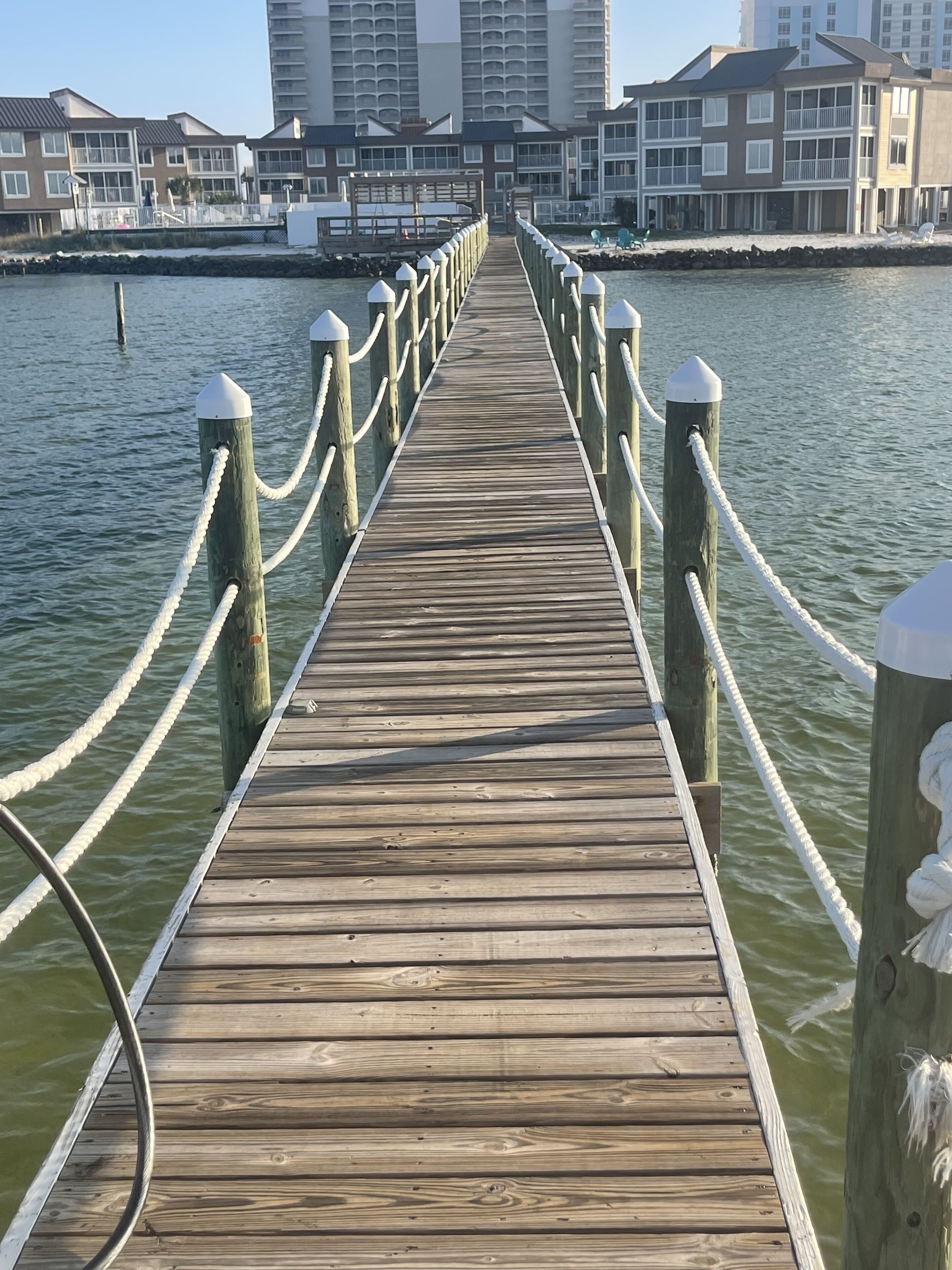 A wooden pier with white ropes and posts, leading to a waterfront area with buildings in the background.
