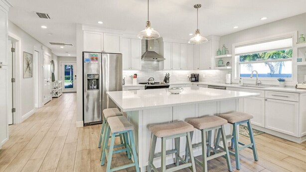 A modern kitchen with white cabinetry, a central island with stools, pendant lighting, and a view of the outdoors through a window.