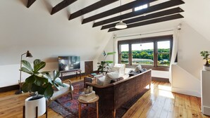 A spacious living room with a high ceiling, wooden beams, a large window with a view of greenery, a sofa, a coffee table, and a television. - The Pearl, NH hidden luxury close to the Quinnipiac River in historic property (New Haven)
