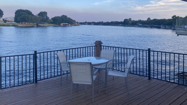 Une terrasse en bois avec une table et des chaises, donnant sur une étendue d’eau calme, avec des bâtiments et des arbres en arrière-plan.