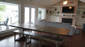 Un salon spacieux avec une grande table en bois, un canapé gris, une cheminée en briques et un téléviseur fixé au mur.