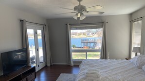 Une chambre à coucher avec un lit, un téléviseur posé sur un meuble en bois, un ventilateur de plafond et une vue sur un lac par la fenêtre.