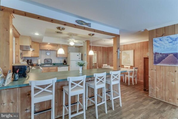 A modern kitchen with wooden cabinets, a central island with bar stools, pendant lighting, and a large window with a view. - Quaint Beach Block Cottage, 100 steps to the beach (Dewey Beach)