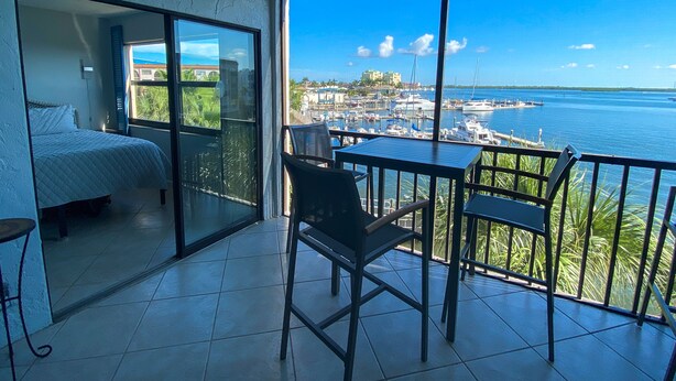 A balcony with a bed, a table, and chairs, overlooking a marina with boats and a distant town. - MarcoView 402 - A Slice of Paradise! Premier Waterfront Location @Anglers Cove! (Marco Island)
