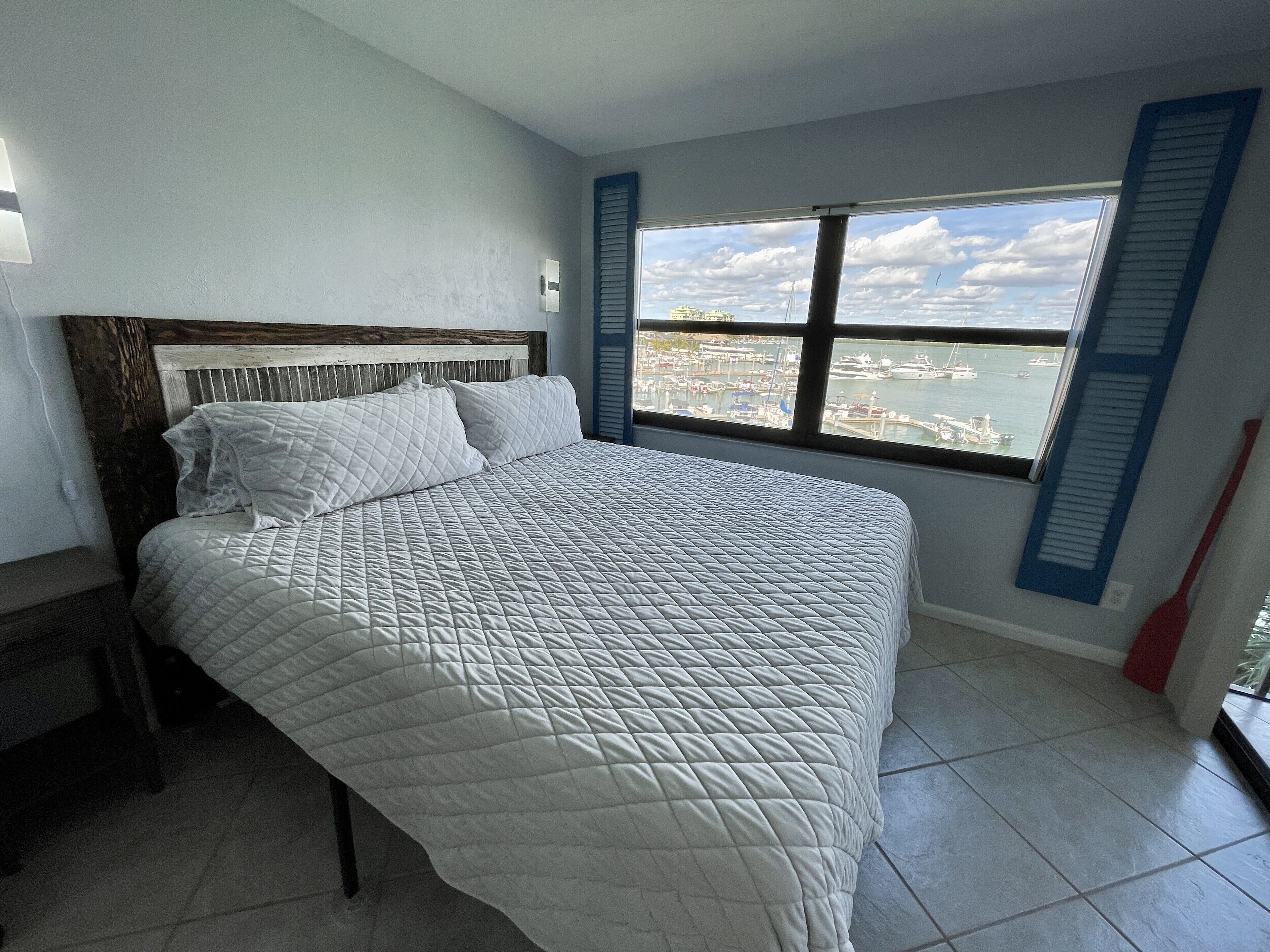 A bedroom with a bed, a window with a view of a marina, and a red broom leaning against the wall.