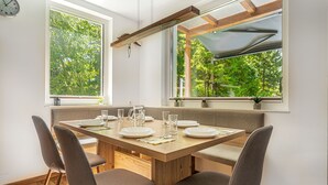 Un coin repas avec une table en bois dressée pour quatre personnes, de grandes fenêtres donnant sur un paysage verdoyant et un ventilateur de plafond.
