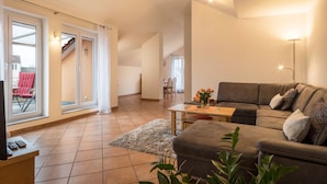 Ein geräumiges Wohnzimmer mit einer Couch, einem Couchtisch und einem Fernseher. Eine Glastür öffnet sich zu einem Außenbereich mit einem roten Sessel.
