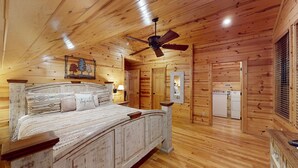 A wooden cabin bedroom with a bed, a ceiling fan, a refrigerator, and a window with blinds.