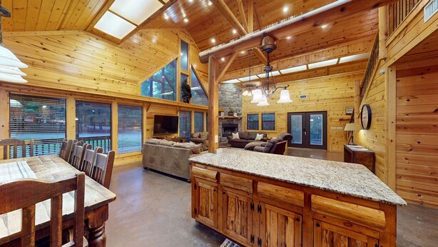 A spacious wooden cabin interior with a dining area, a living room with a fireplace, and a large window overlooking the outdoors.