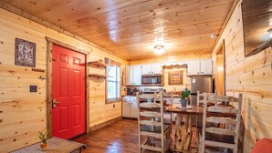 Interior de una cabaña de madera con una puerta roja, una mesa de comedor con sillas, una zona de cocina con electrodomésticos y una mesita con una planta.