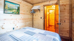 Un dormitorio de cabaña de madera con una cama, un cuadro y una puerta que da a una habitación iluminada cálidamente.