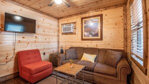 Interior acogedor de una cabaña con techo de madera, ventilador de techo, televisor de pantalla plana, un sillón rojo, un sofá marrón con un cojín que dice 'lugar feliz' y una mesita con una planta.
