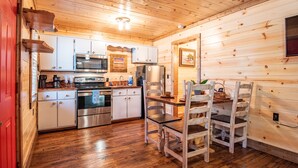 Interior de una cabaña de madera con cocina que cuenta con armarios blancos, electrodomésticos de acero inoxidable y comedor con sillas y mesa de madera.