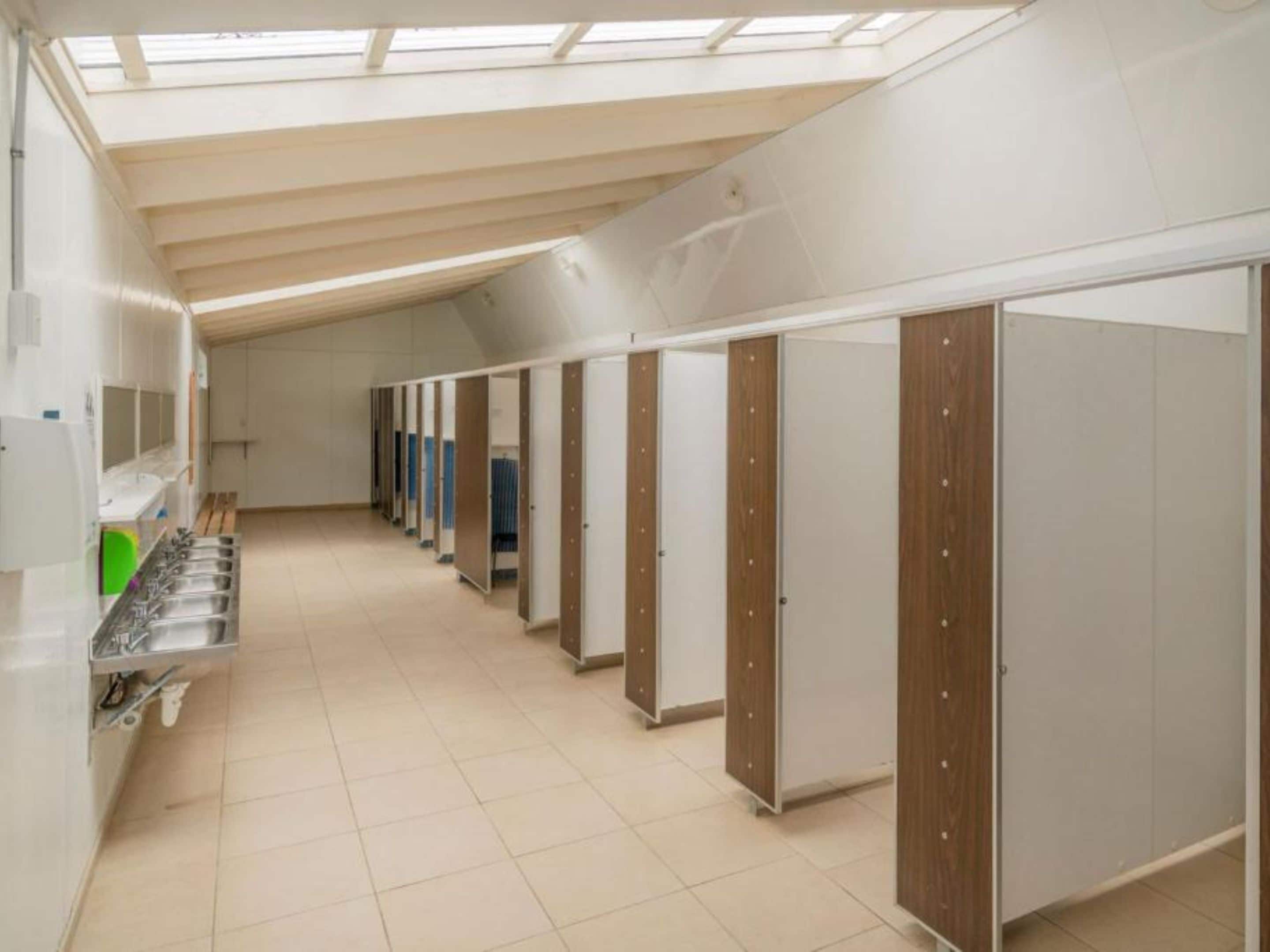 Une rangée de chambres individuelles dotées de portes en bois, d’un puits de lumière et d’un espace lavabo équipé de sèche-mains.