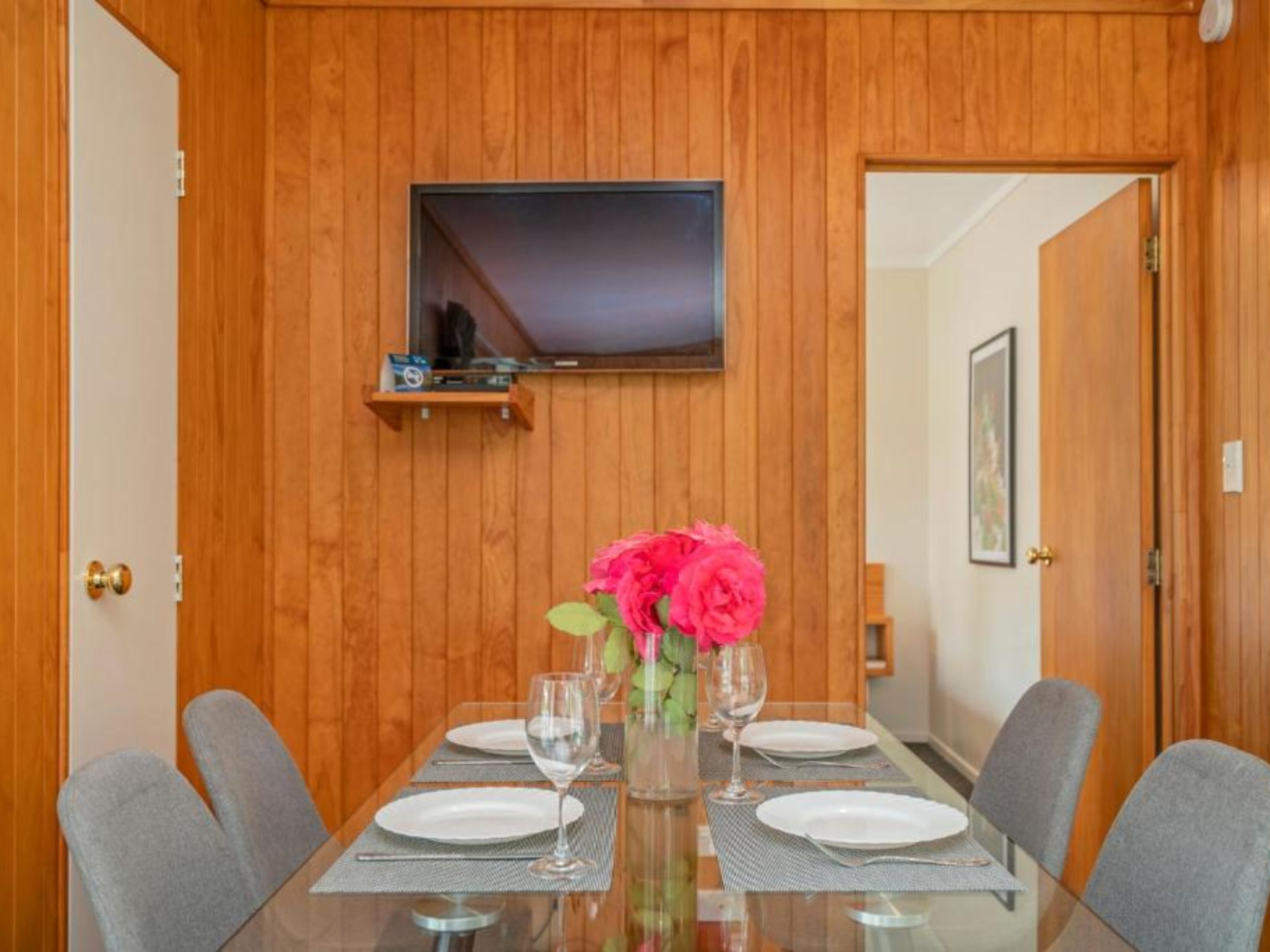 A dining area with a wooden paneled wall, a flat-screen TV mounted above a shelf, and a table set for two with a vase of flowers.