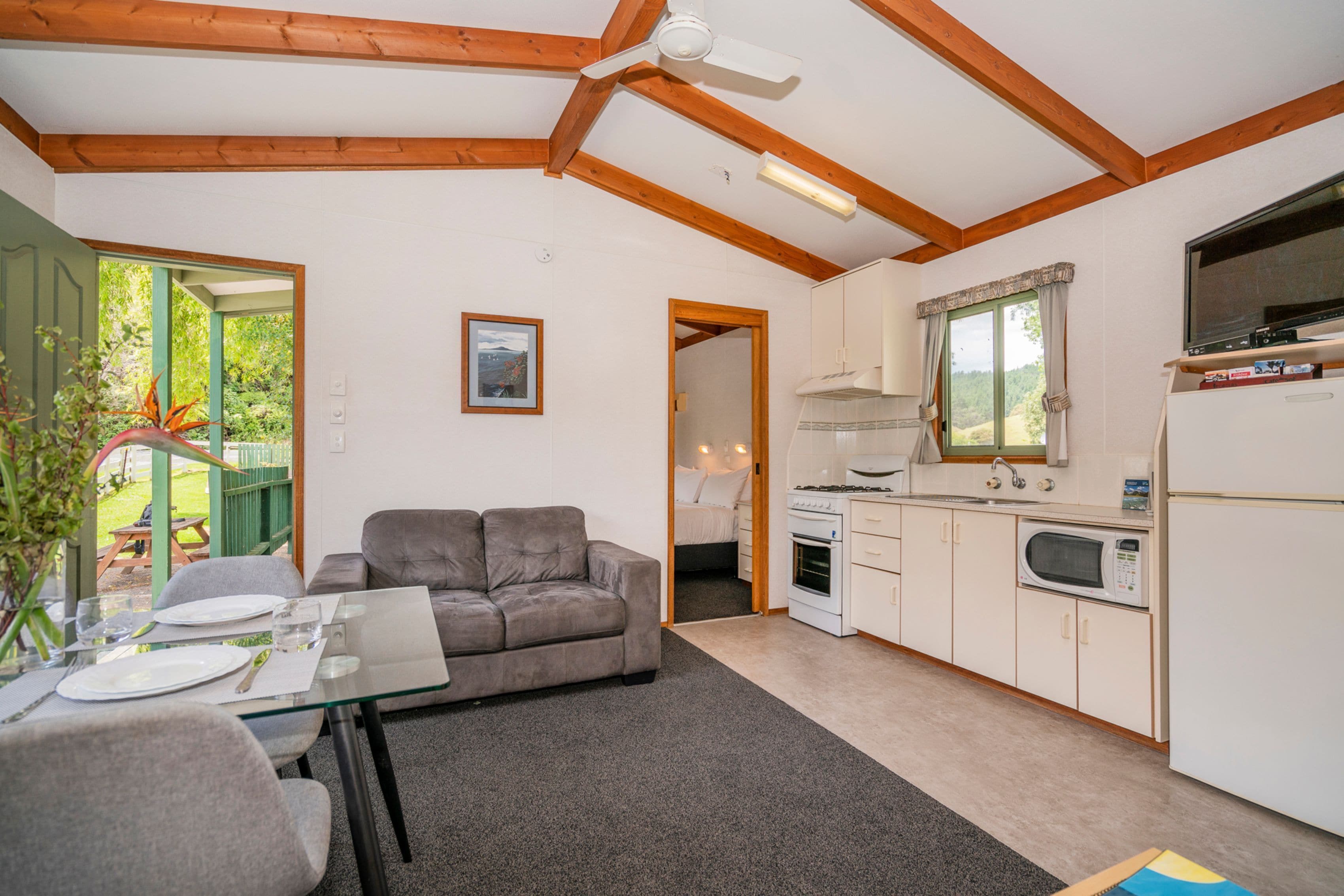 Un espacio habitable compacto con kitchenette, una zona de comedor con mesa de vidrio, un sofá gris y un ventilador de techo.