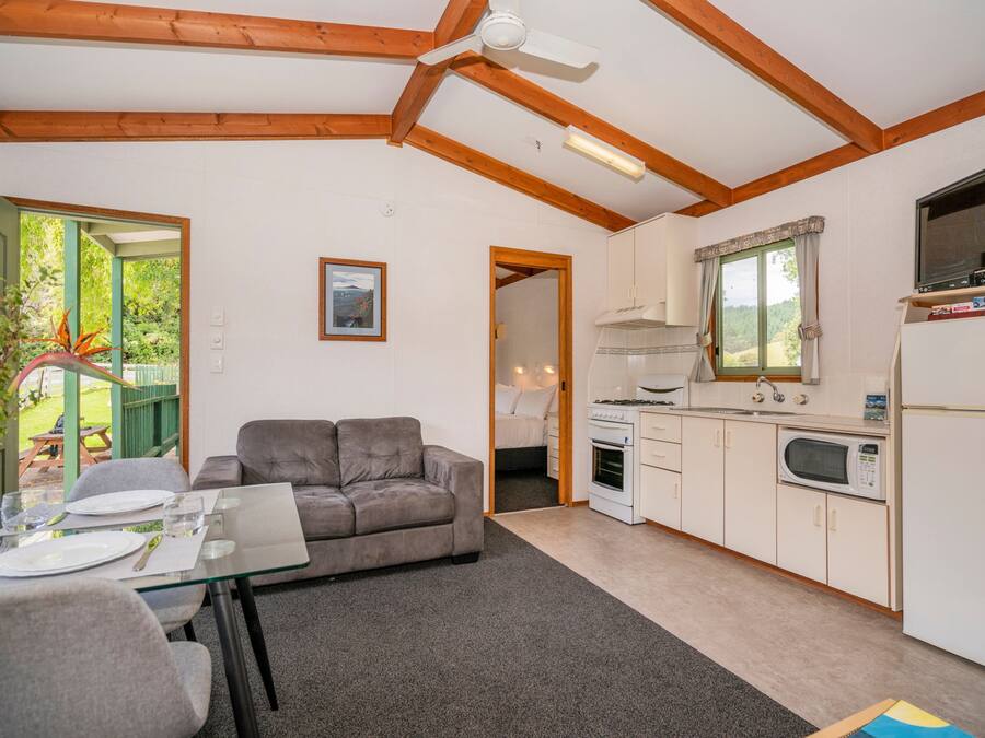 Un espacio habitable compacto con kitchenette, una zona de comedor con mesa de vidrio, un sofá gris y un ventilador de techo.