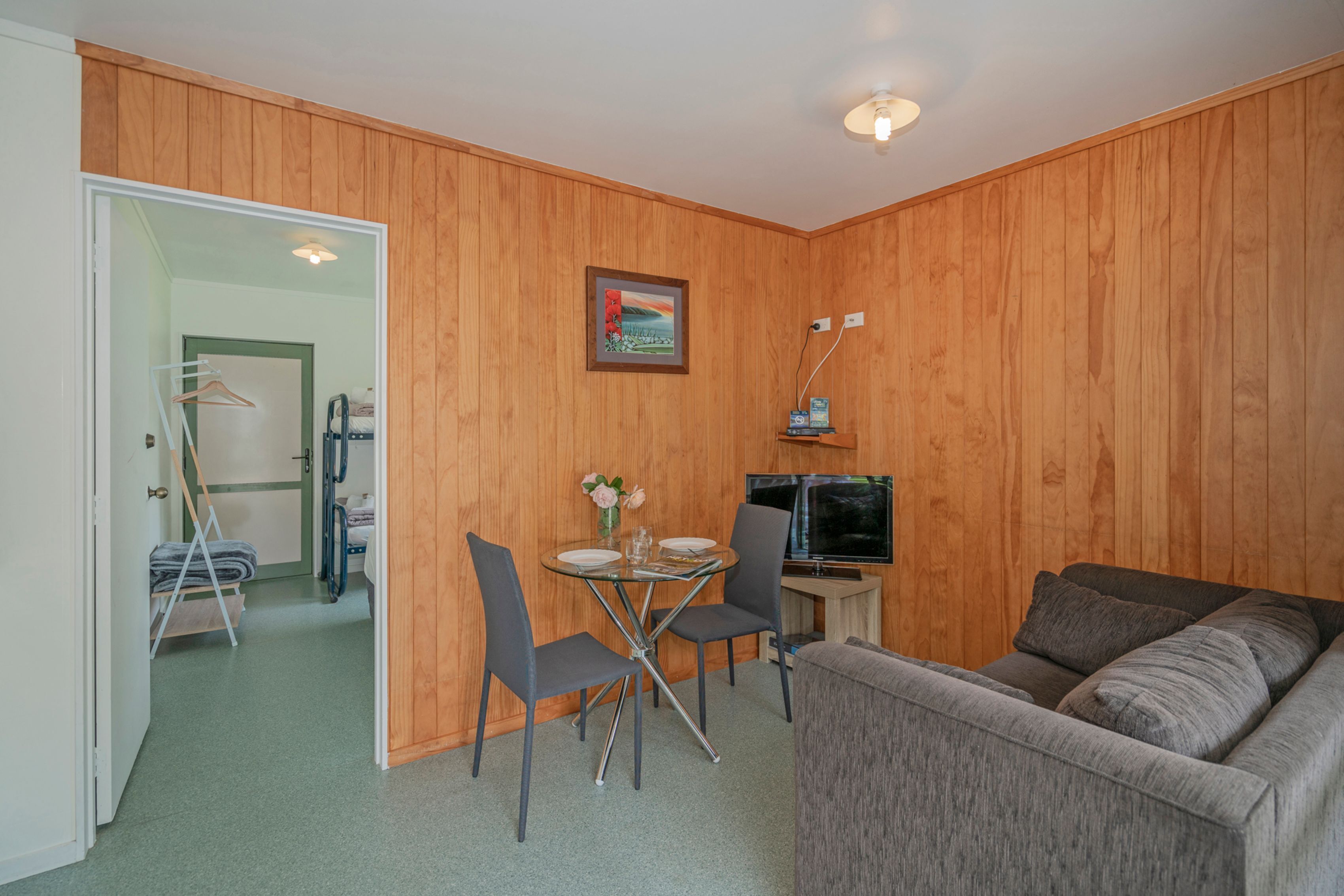 Un espace de vie compact avec un mur en bois, un canapé gris, une petite table à manger avec des chaises, un téléviseur et un tableau accroché au mur.